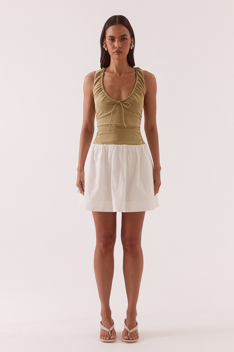 Model wearing a mixed fabric mini dress with a stretchy sleeveless bodice, deep scoop neckline with adjustable drawstring, racer back, gathered detailing through the bodice and skirt, side pockets, and a fully lined silhouette.