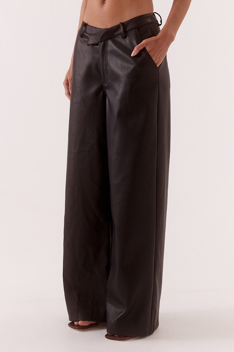 Model wearing black faux leather wide leg pants with angled waistband, fly front closure, and functional pockets, styled in a high-waisted tailored fit.