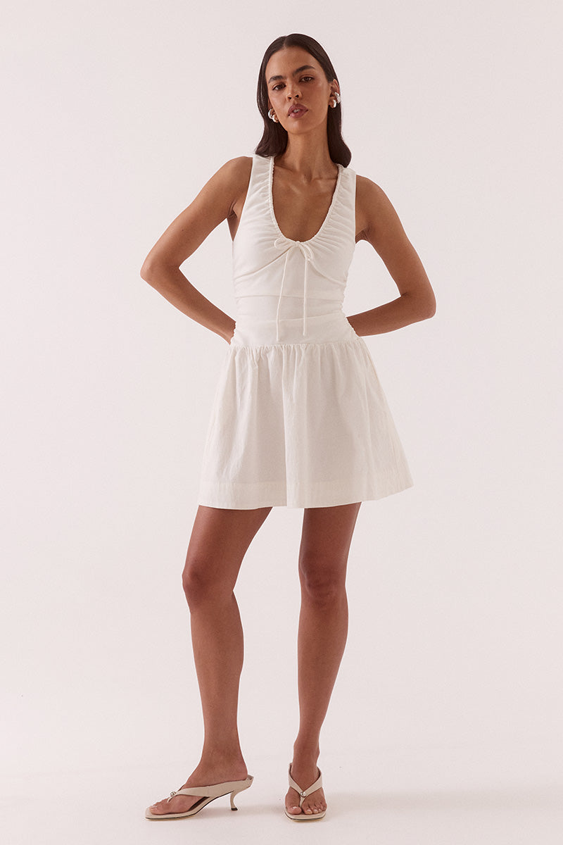 Model wearing a mixed fabric mini dress with a stretchy sleeveless bodice, deep scoop neckline with adjustable drawstring, racer back, gathered detailing through the bodice and skirt, side pockets, and a fully lined silhouette.