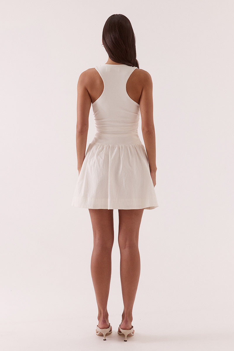 Model wearing a mixed fabric mini dress with a stretchy sleeveless bodice, deep scoop neckline with adjustable drawstring, racer back, gathered detailing through the bodice and skirt, side pockets, and a fully lined silhouette.