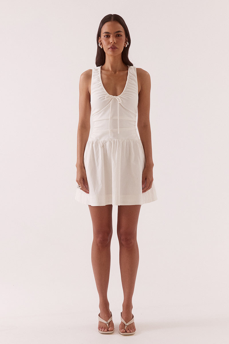 Model wearing a mixed fabric mini dress with a stretchy sleeveless bodice, deep scoop neckline with adjustable drawstring, racer back, gathered detailing through the bodice and skirt, side pockets, and a fully lined silhouette.