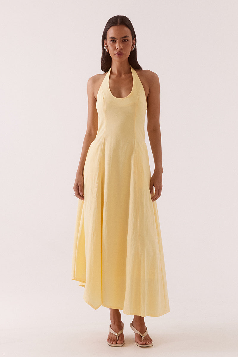 Model wearing a cotton blend denim halter neck maxi dress featuring a scoop neckline, fitted bodice, full skirt with an asymmetric hem, front godet insert, hidden side seam pockets, and a fully lined silhouette.