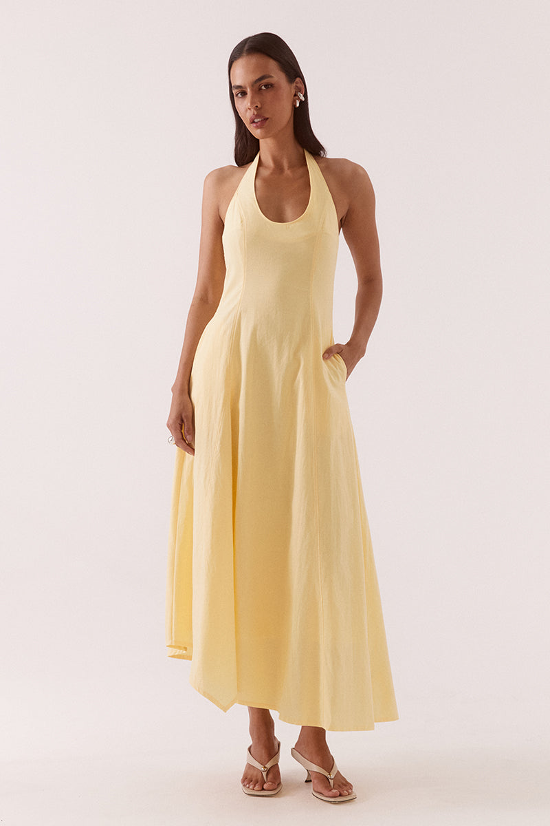 Model wearing a cotton blend denim halter neck maxi dress featuring a scoop neckline, fitted bodice, full skirt with an asymmetric hem, front godet insert, hidden side seam pockets, and a fully lined silhouette.