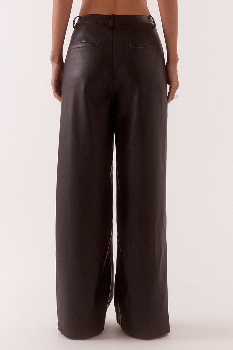 Model wearing black faux leather wide leg pants with angled waistband, fly front closure, and functional pockets, styled in a high-waisted tailored fit.